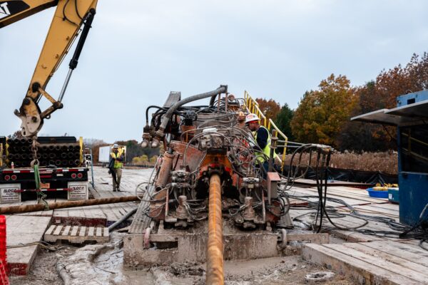 Workers operate drills at muddy site with trees and equipment.