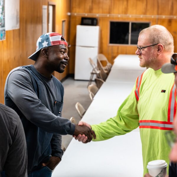 Two men shake hands in a meeting room, others watching.