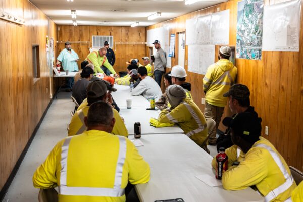 Workers in yellow safety gear meet in a wood-paneled room.