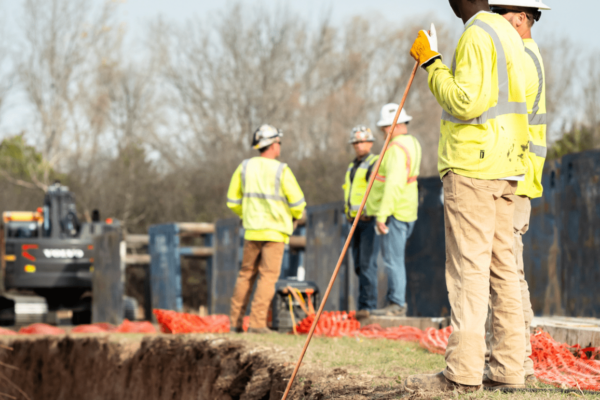 Construction workers in safety gear stand by trench and equipment.