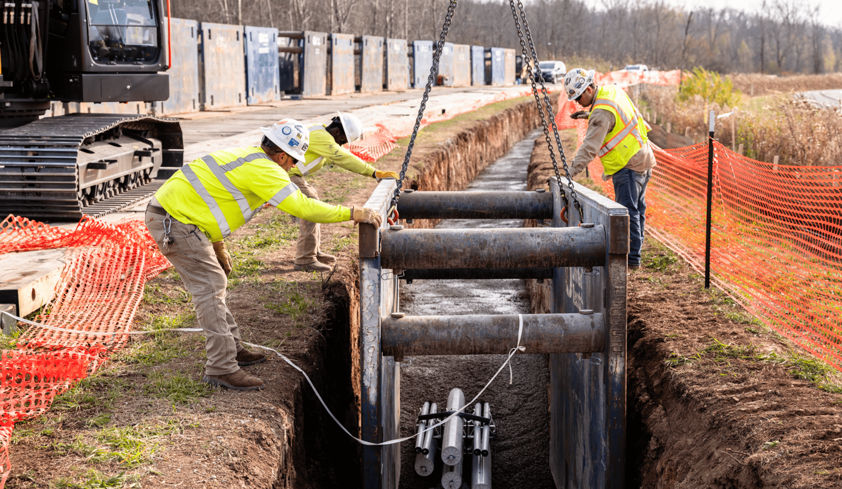 Two construction workers install a trench box with crane; pipes exposed.