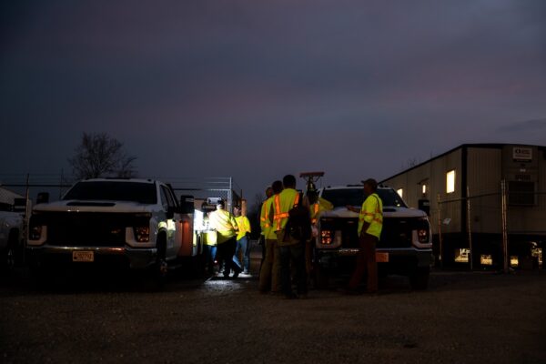 Workers in vests talk between trucks in dim parking lot.
