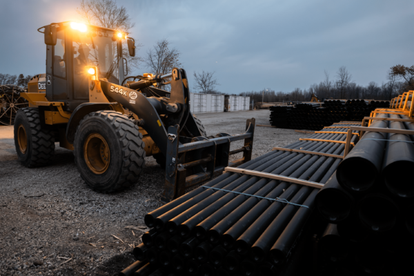 Yellow front loader with headlights by black pipes at dusk.