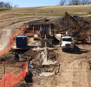 Excavator, truck, and workers install utility pipeline along dirt road in Jefferson City, Missouri.