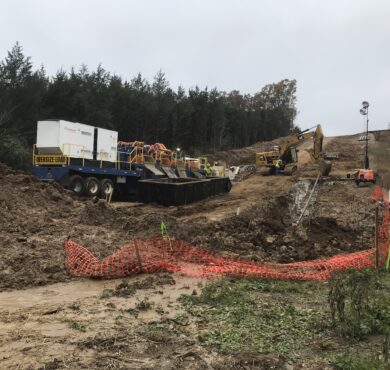 Excavators and trucks work on muddy site near forest, fenced.