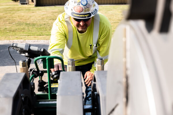 Worker in hard hat operates machinery near yellow and brown building.
