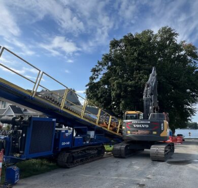 Volvo excavator beside blue conveyor on lakeside residential street.