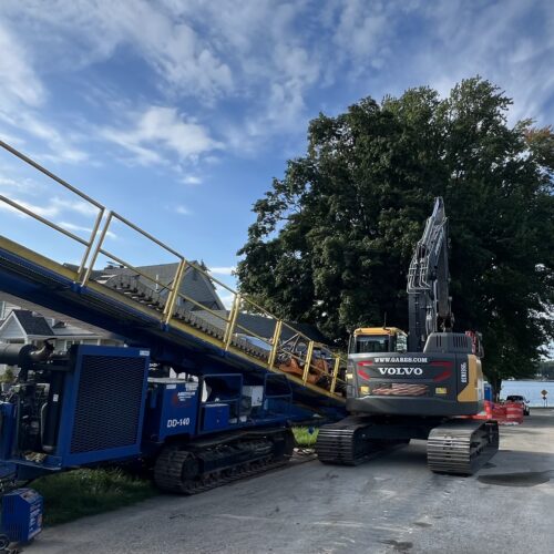 Volvo excavator beside blue conveyor on lakeside residential street.