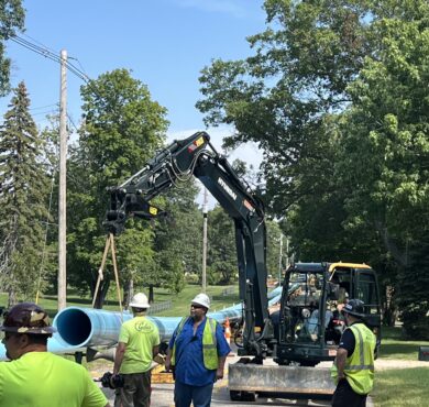 Workers use excavator to place blue utility pipe on street.