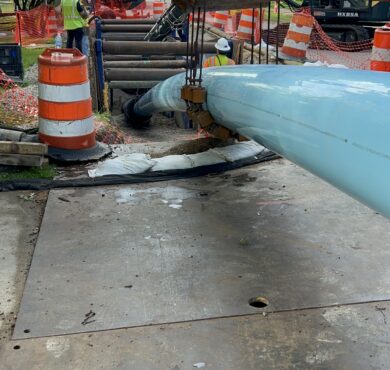 Workers install a large blue pipe underground amid safety barriers.