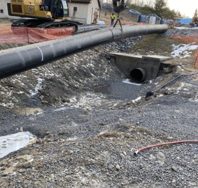 Excavator places pipeline over culvert; gravel, snow, cones visible.