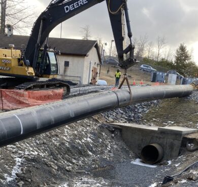 Excavator places pipeline over culvert; gravel, snow, cones visible.