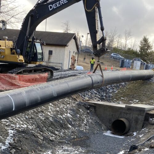 Excavator places pipeline over culvert; gravel, snow, cones visible.