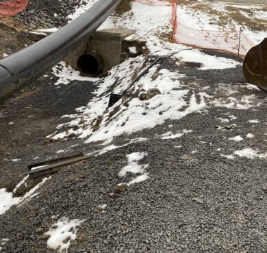 Construction site: black pipe over culvert, snow, gravel, excavator.