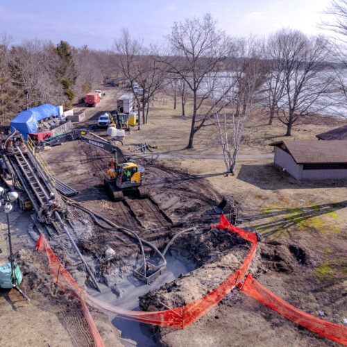 Horizontal directional drilling construction site with machinery, mud, barriers, lake, and bare trees at Dunes State Park