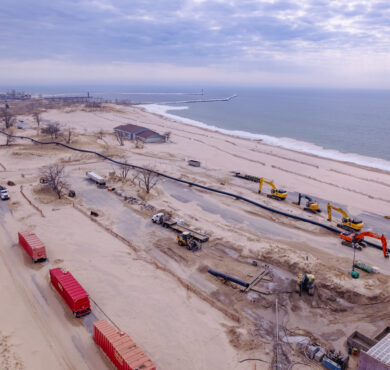 Aerial view: construction vehicles on sandy beach at Dunes State Park under cloudy sky.