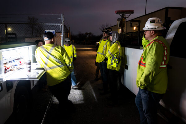 Construction workers in reflective gear discuss equipment by lit truck.