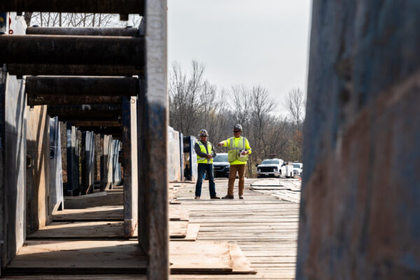 Two workers in safety gear stand on platform, trench boxes in foreground.