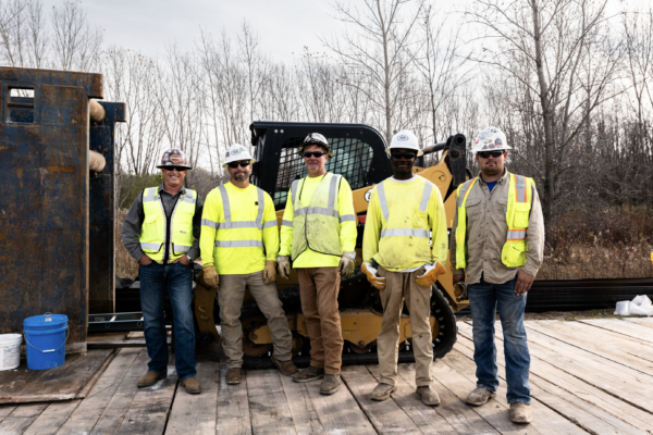 Five construction workers wearing safety attire stand in front of a trench box and skid steer.