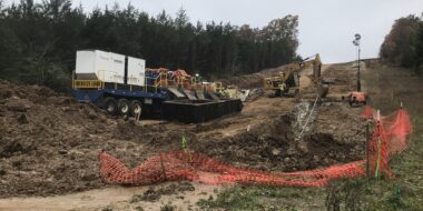 Underground utility construction site with machinery, trucks, trees, and orange fencing