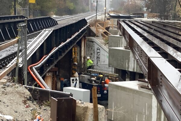 Workers in safety gear operate equipment to install fiber-optic cable under demolished and new bridge.