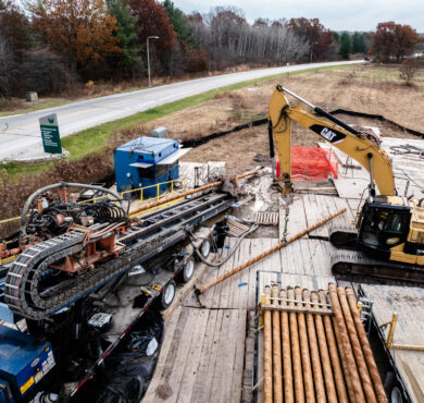 Construction site with excavator, drilling equipment, wood, and trees.