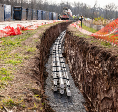 Utility pipes in trench, orange fencing and machinery behind.