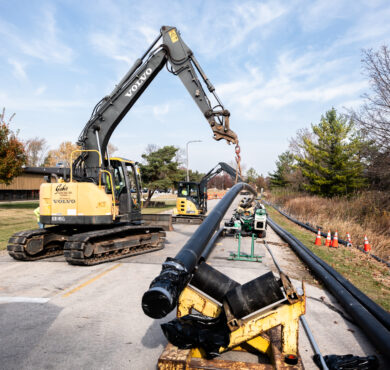 Excavator lifts black pipe near road, cones, and equipment.