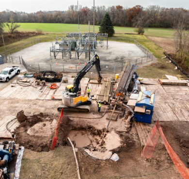 Excavator and workers operate on muddy site near fenced high-voltage substation.