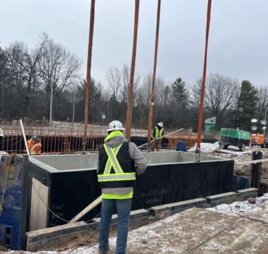 Workers install high-voltage utility concrete structure at snowy, tree-surrounded site.