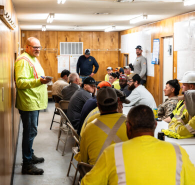 Construction workers in safety gear listen to safety presenter with notepad.