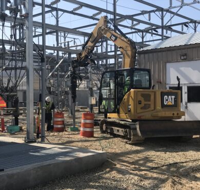 Yellow CAT excavator works at electrical substation with workers and barrels.
