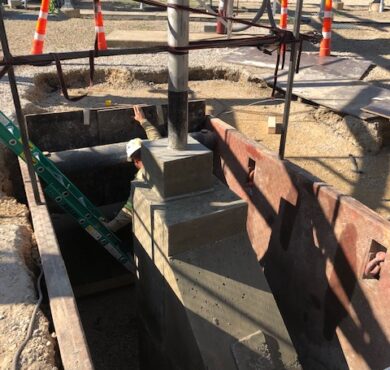 Construction worker in trench box with pipes, equipment, and safety cones.