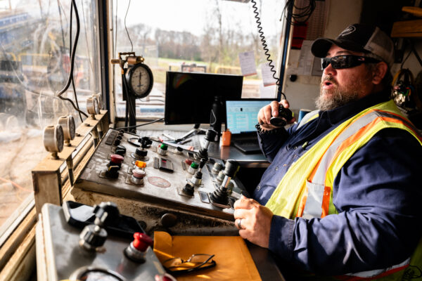 Man in safety vest operates machinery, speaks into radio, laptop nearby.