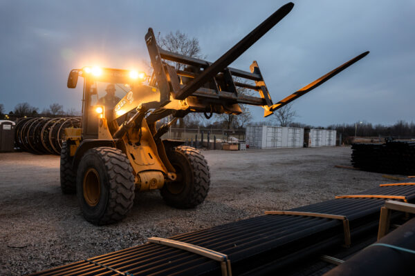 Forklift lifts materials at dusk, headlights on, pipes behind.