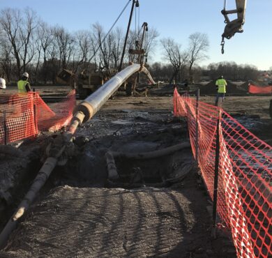 Workers install large utility pipeline with machinery and orange netting.