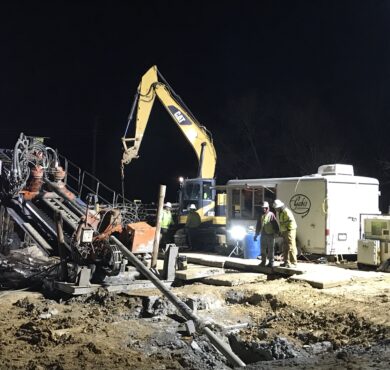 Construction workers use heavy machinery and horizontal directional drill at night under bright lights.