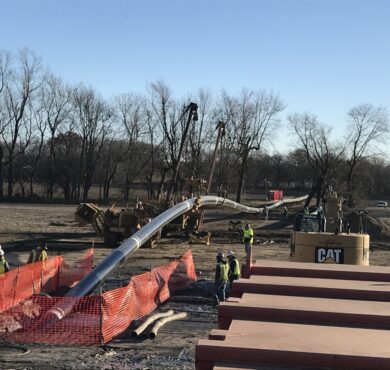 Workers install large pipeline with cranes, fenced site, trees nearby.