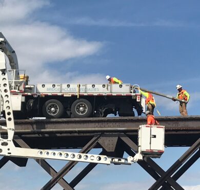 Three workers remove fiber-optic cable from a railroad bridge on a truck; one operates lift.