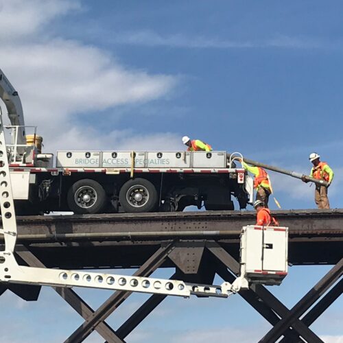 Three workers remove fiber-optic cable from a railroad bridge on a truck; one operates lift.