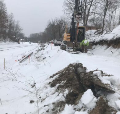 Worker excavates trench with HDD by snowy roadside near train tracks. Winter utility installation in Merrimac, Wisconsin.