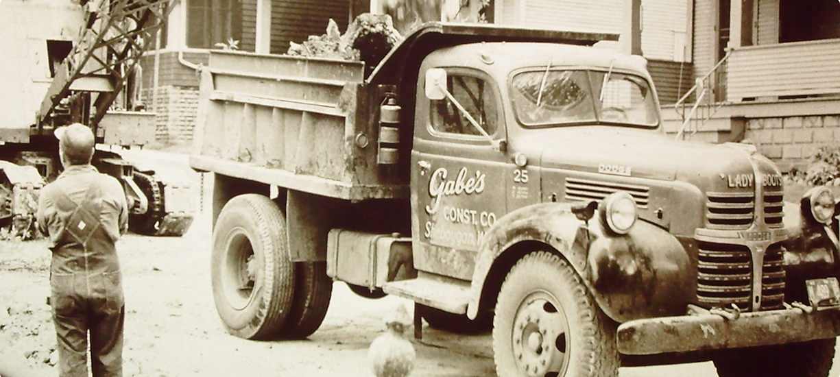 Vintage Gabes Const. Co. dump truck parked by worker, house behind.