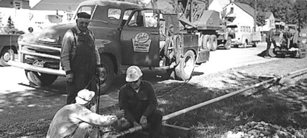 Black-and-white historical photo showing three men working on a pipe near parked utility trucks in 1960