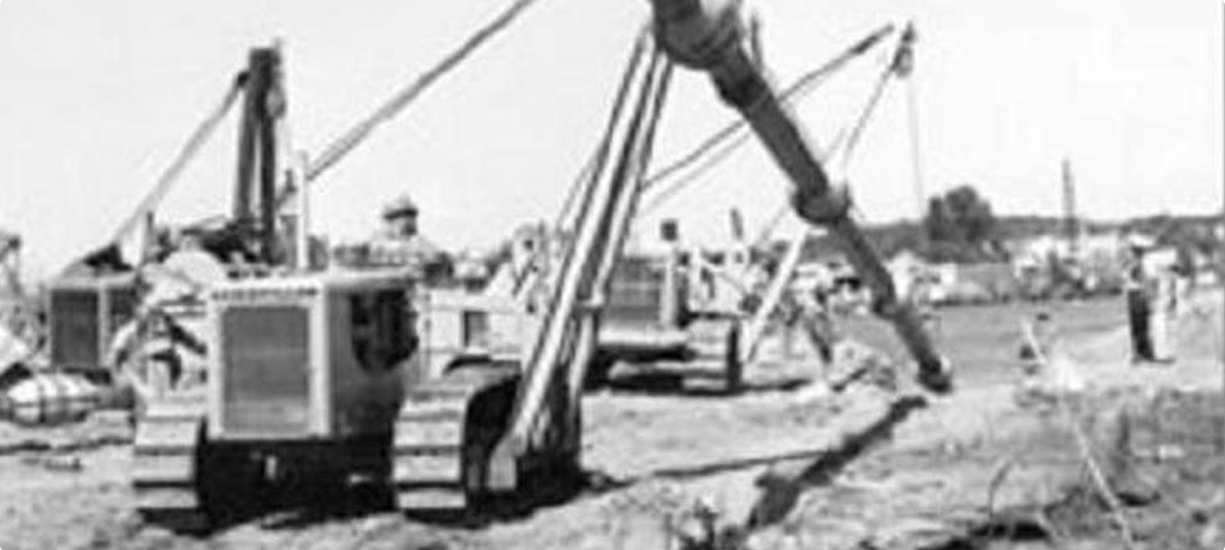 Workers operate heavy machines, laying pipes on dirt path.