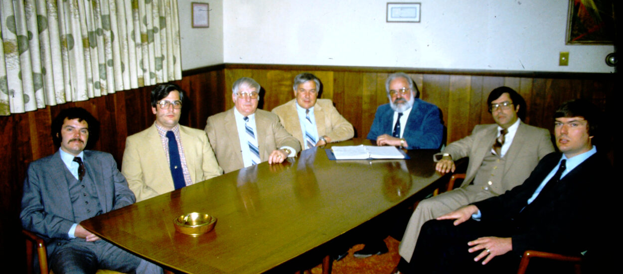 Seven suited men, leaders of Gabe's, sit around a table in wood-paneled office.