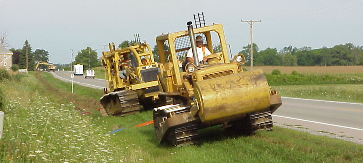 Two yellow bulldozers travel beside a rural highway.