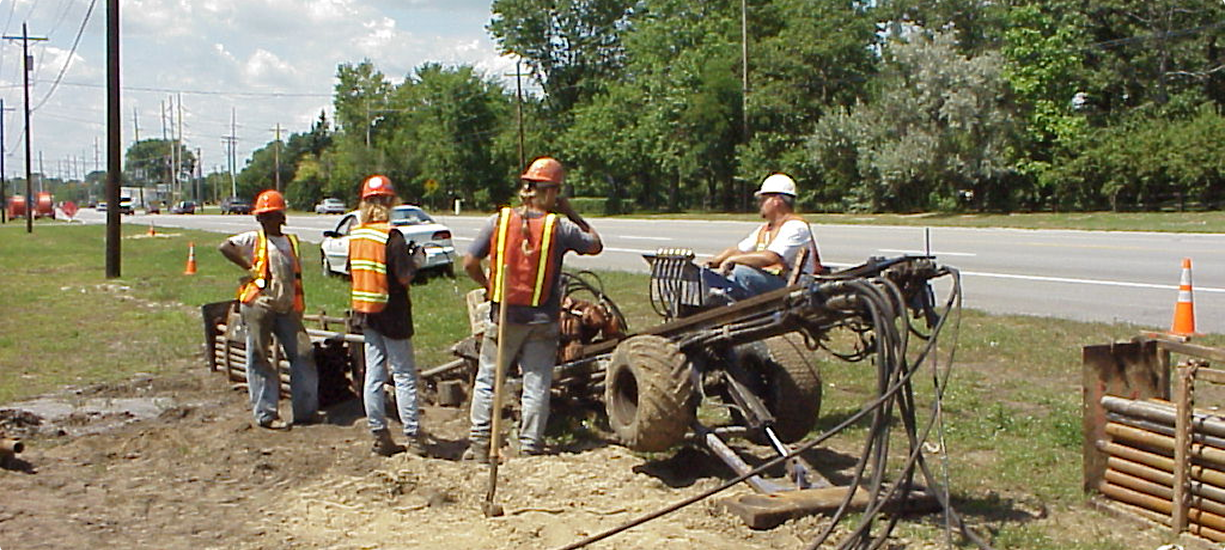 Four workers in safety gear near equipment and roadside cones.