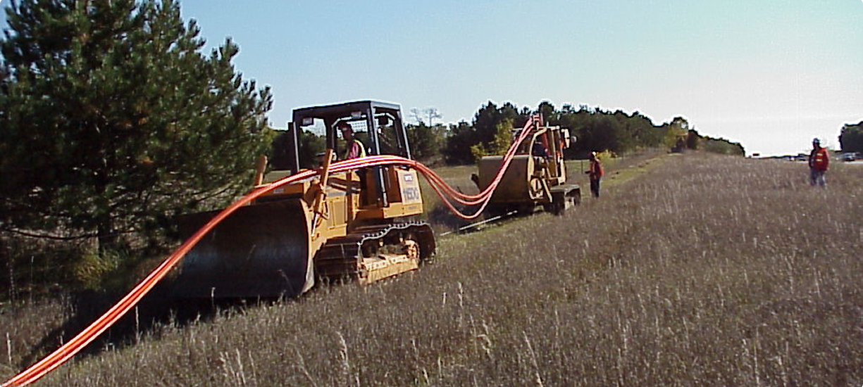 Bulldozer pulls orange cables across field; workers, trees nearby.
