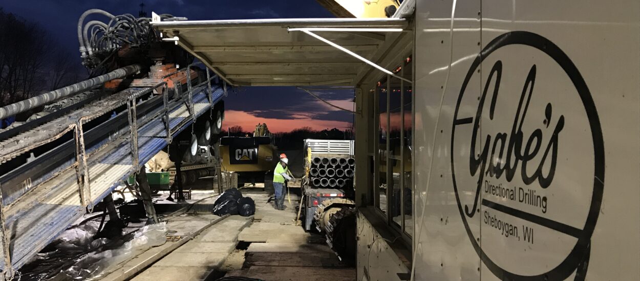 Construction site at dusk: workers, equipment, Gabe’s Drilling trailer.