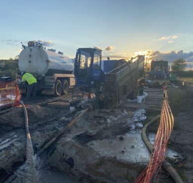 Workers operate machinery and tanker at muddy, fenced sunset wetlands construction site.